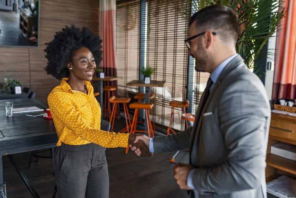 Two people wearing business clothing are standing and shaking hands, with small coffee tables and chairs in the background. This image represents a job developer connecting with an employer.