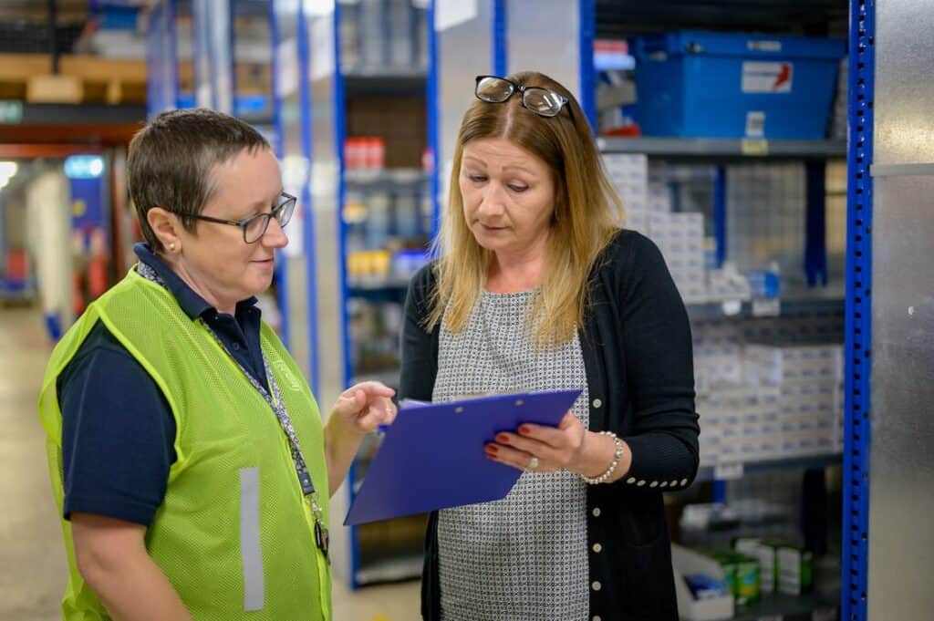 In a warehouse, an employer and employment practitioner review a document on a clipboard. This image represents a job-site analysis (JSA), one of the tools discussed in the Supported Employment Essentials program.