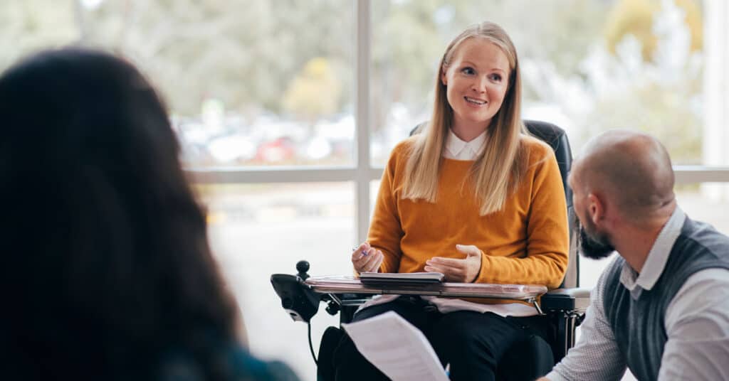 Photo: Person in business attire in wheelchair with a tablet speaking to other people in a workplace