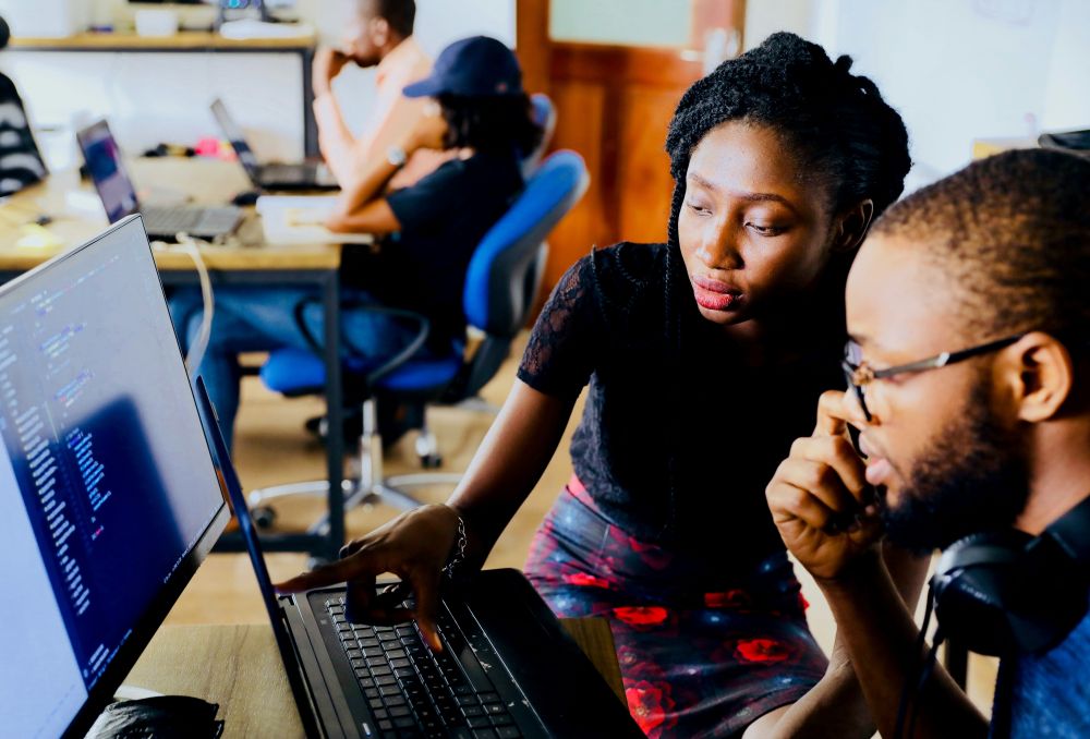 A mentor and a mentee, both with medium skin tone, look at a computer monitor in a workplace.